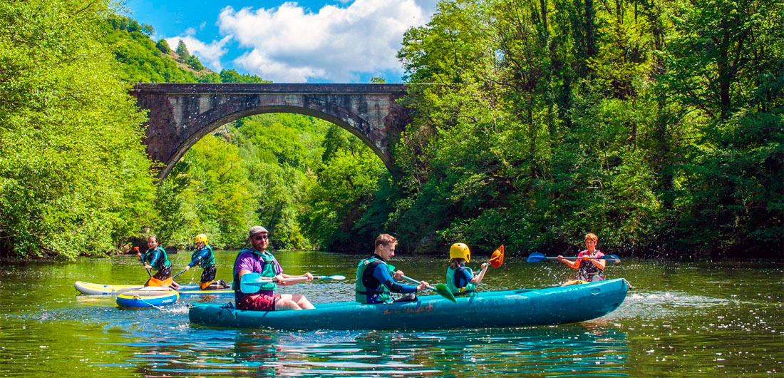 Famille en canoë sur la rivière Lot près du Domaine de La Châtaigneraie, entourée de nature et d’un pont de pierre typique du Cantal
